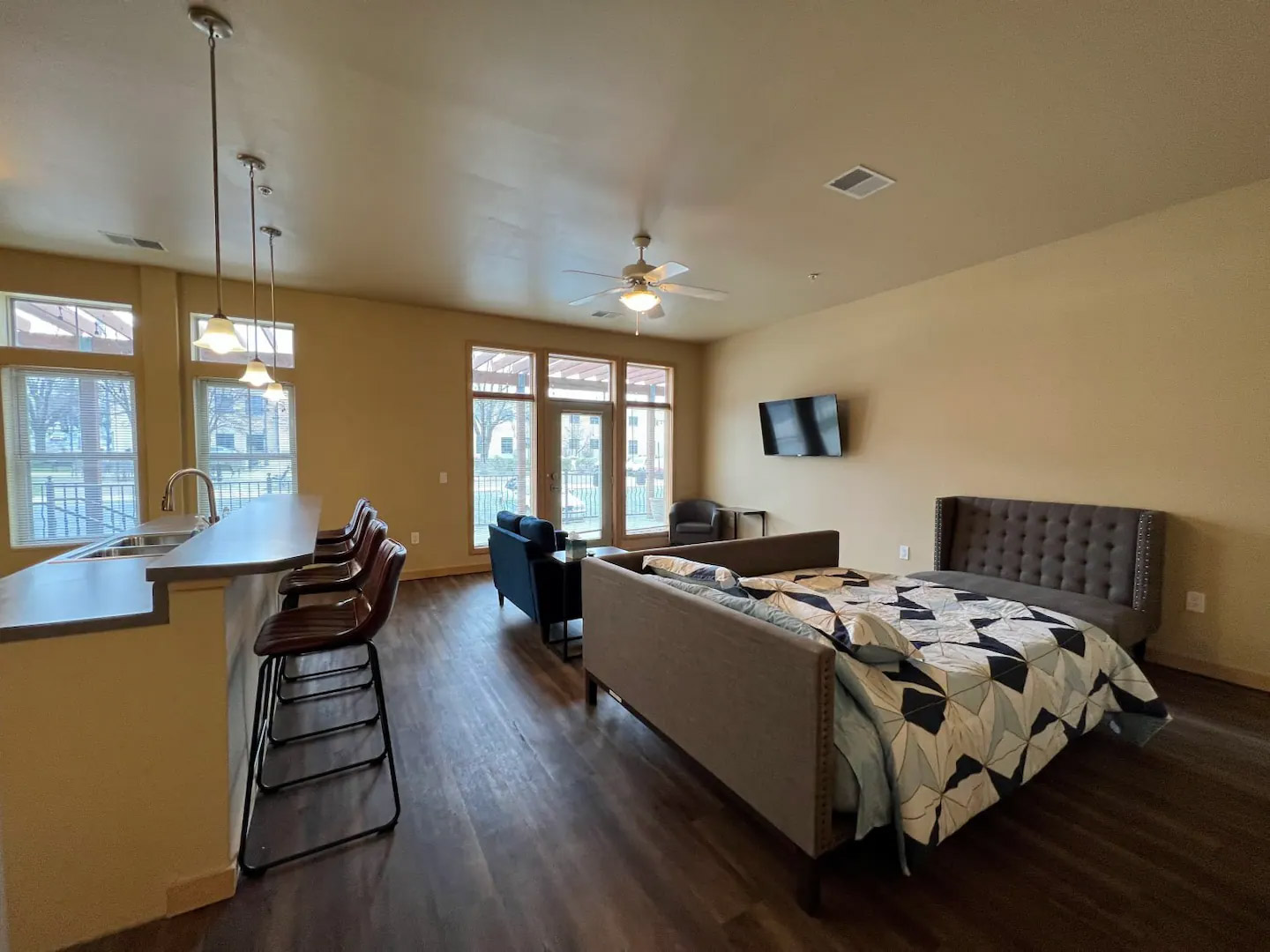 A studio apartment with a double bed, sofa, mounted TV, dining area with bar stools, and a kitchen counter. The room features wood flooring and large windows.