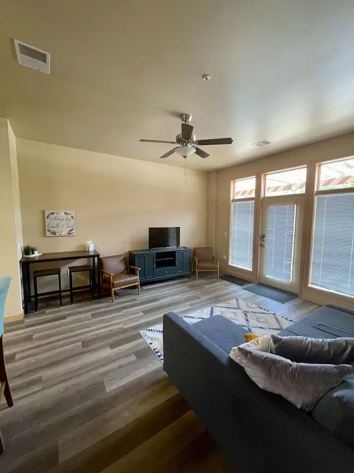 A living room with a ceiling fan, TV on a stand, small side table, chairs, large windows, and a couch with cushions and a blanket. The floor has a mix of wood and light-colored carpet.