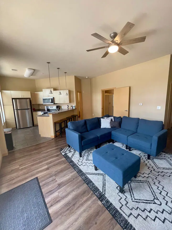 Open-plan living room and kitchen area featuring a blue sectional sofa with matching ottoman, a patterned rug, stainless steel kitchen appliances, an island with barstools, and a ceiling fan.