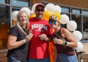 Three adults stand outside in front of white and yellow balloons, smiling and holding drinks. One wears a red Wisconsin Badgers shirt and cap, and all have event wristbands.