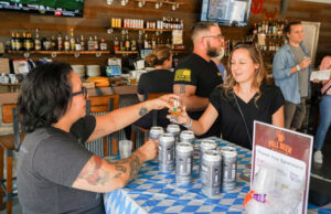 Two people clink shot glasses at a table with beer cans inside a bar, while others stand and socialize in the background.