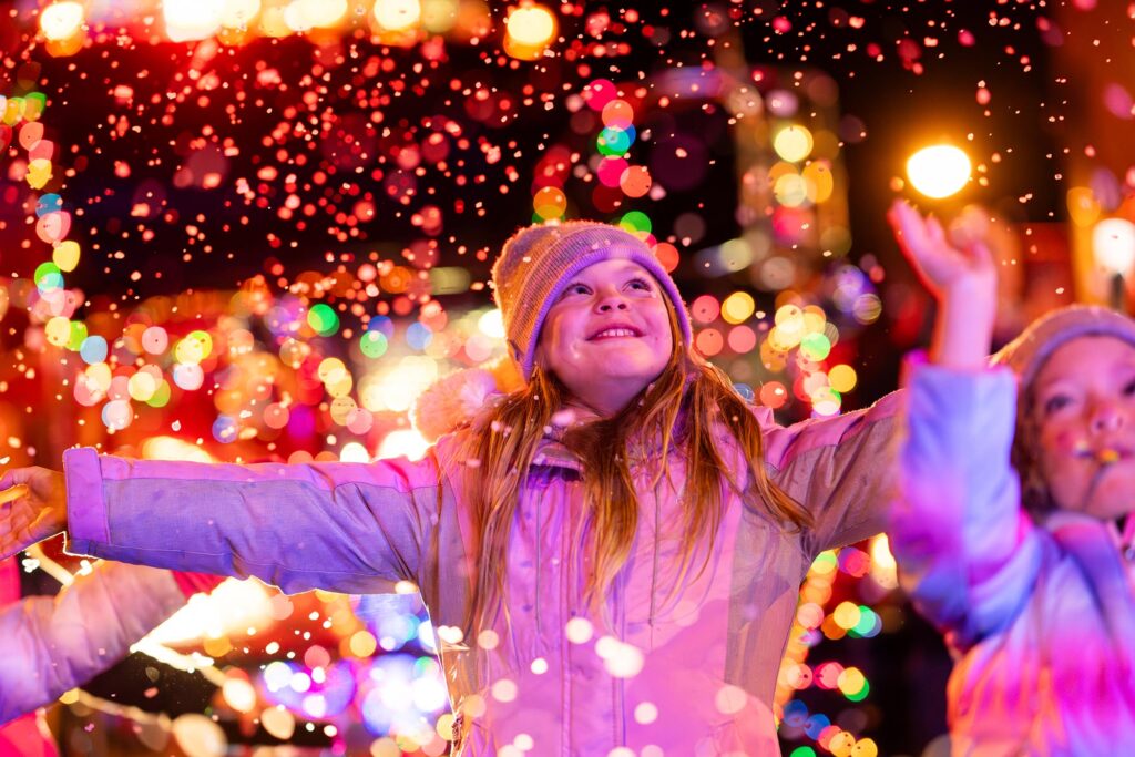 A young girl in a coat and beanie smiles with arms outstretched while surrounded by festive lights and falling confetti.