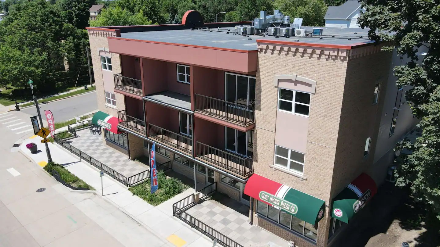 A three-story building with commercial spaces on the ground floor featuring a pizza restaurant with red and green awnings, and balconies on the upper floors. Residential area and trees in the background.