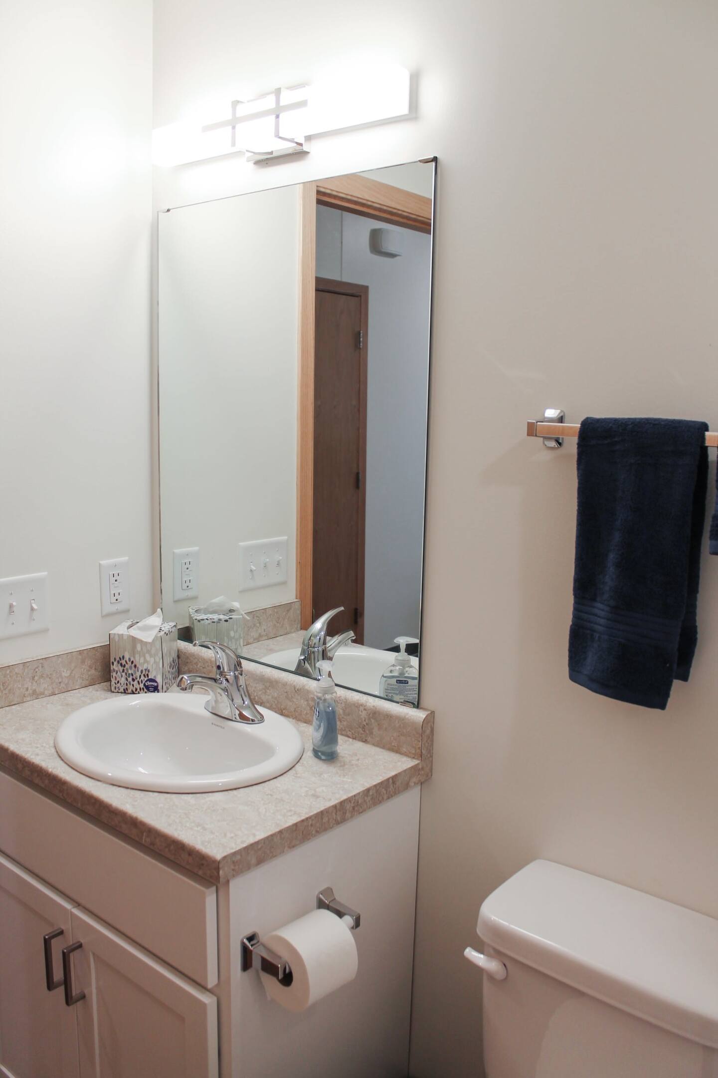 A small bathroom with a sink, mirror, light fixture, countertop, towel rack with a dark blue towel, and a toilet.