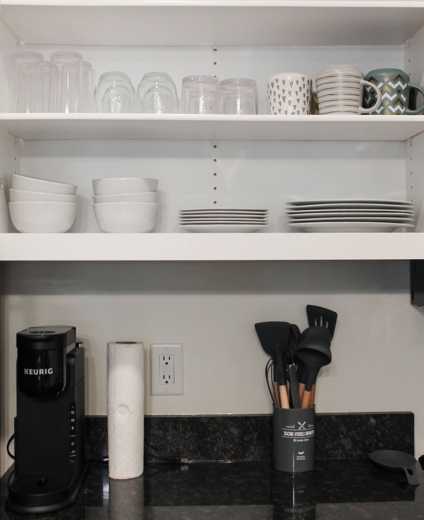 A kitchen shelf with various cups, bowls, and plates neatly arranged. Below, a counter holds a Keurig coffee maker, paper towel roll, and a container with kitchen utensils.