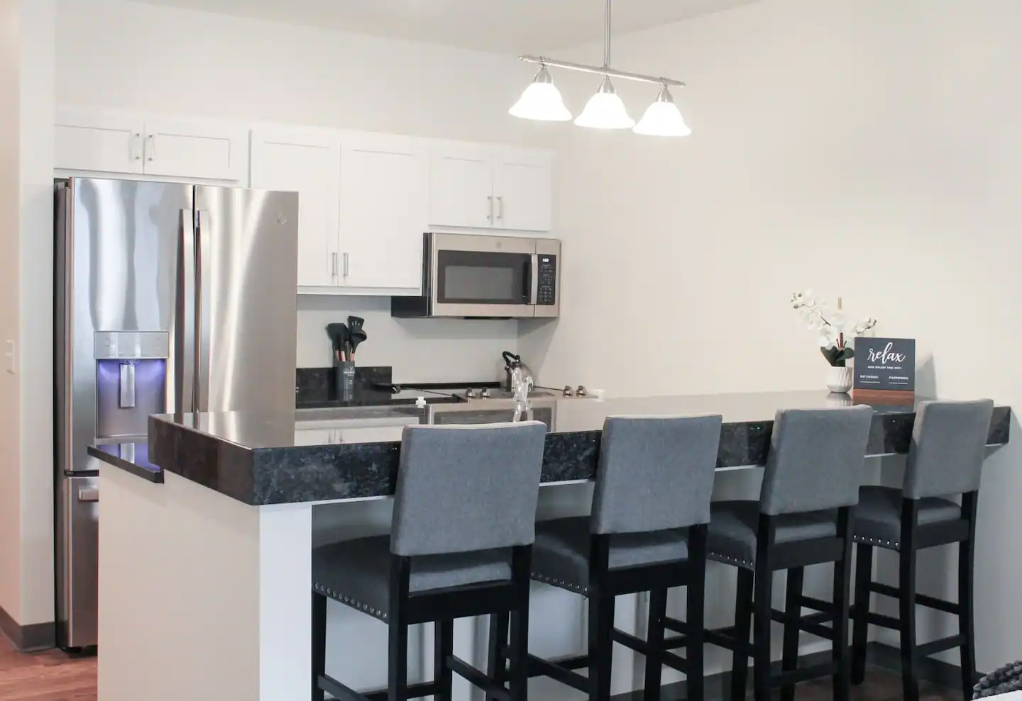 A modern kitchen with a stainless steel refrigerator, microwave, and stove. There is a black granite countertop with four gray bar stools and a three-light pendant hanging above.