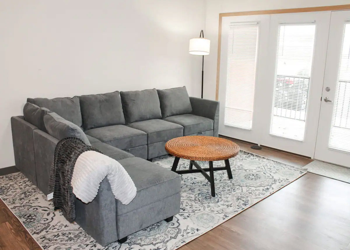 A grey sectional sofa with a throw blanket sits on a patterned rug in a bright living room. A round woven coffee table is in front of the sofa, and a floor lamp is in the corner near the glass door.