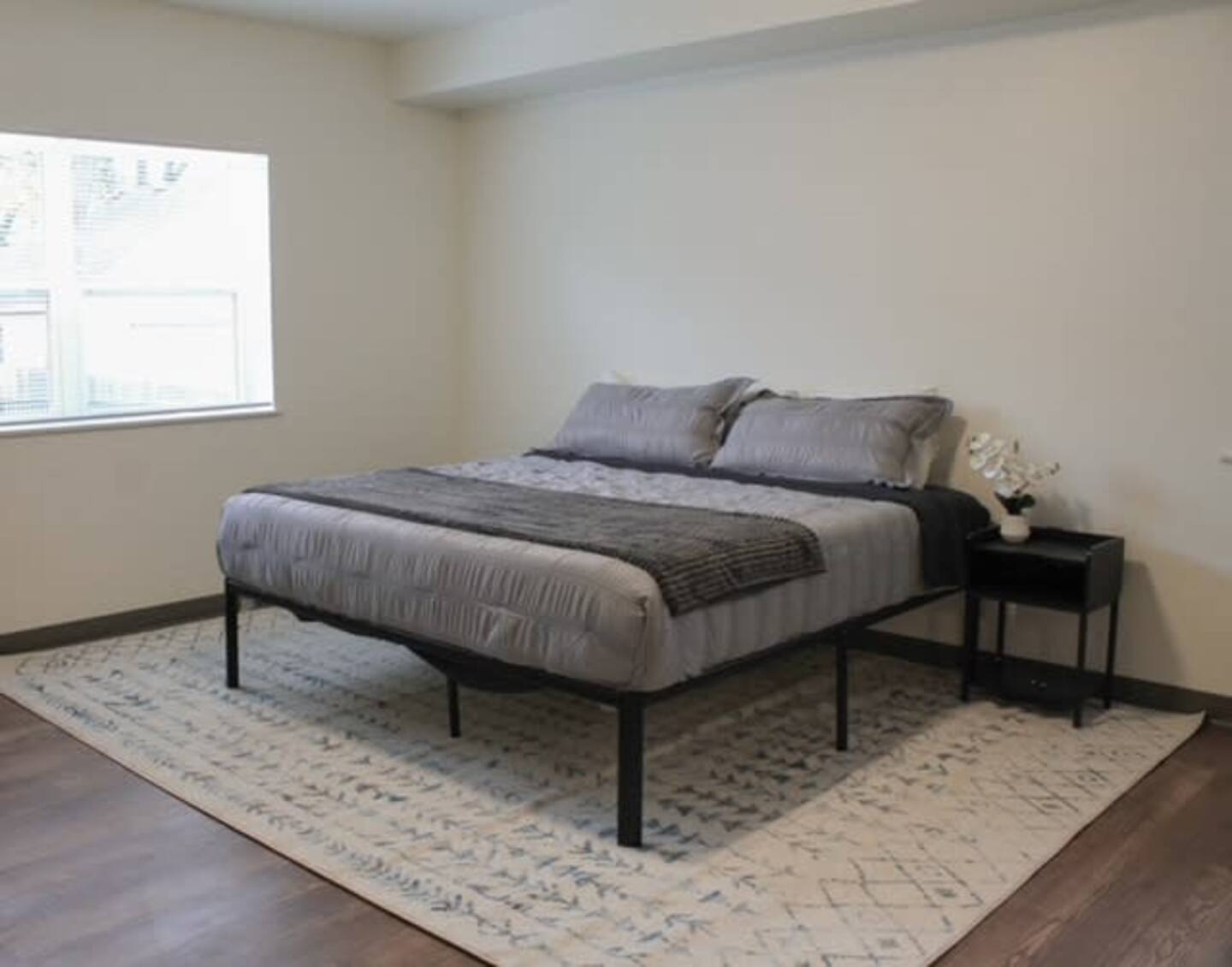 A minimalist bedroom with a neatly made bed, gray bedding, a black nightstand with decorative items, and a window with white blinds.