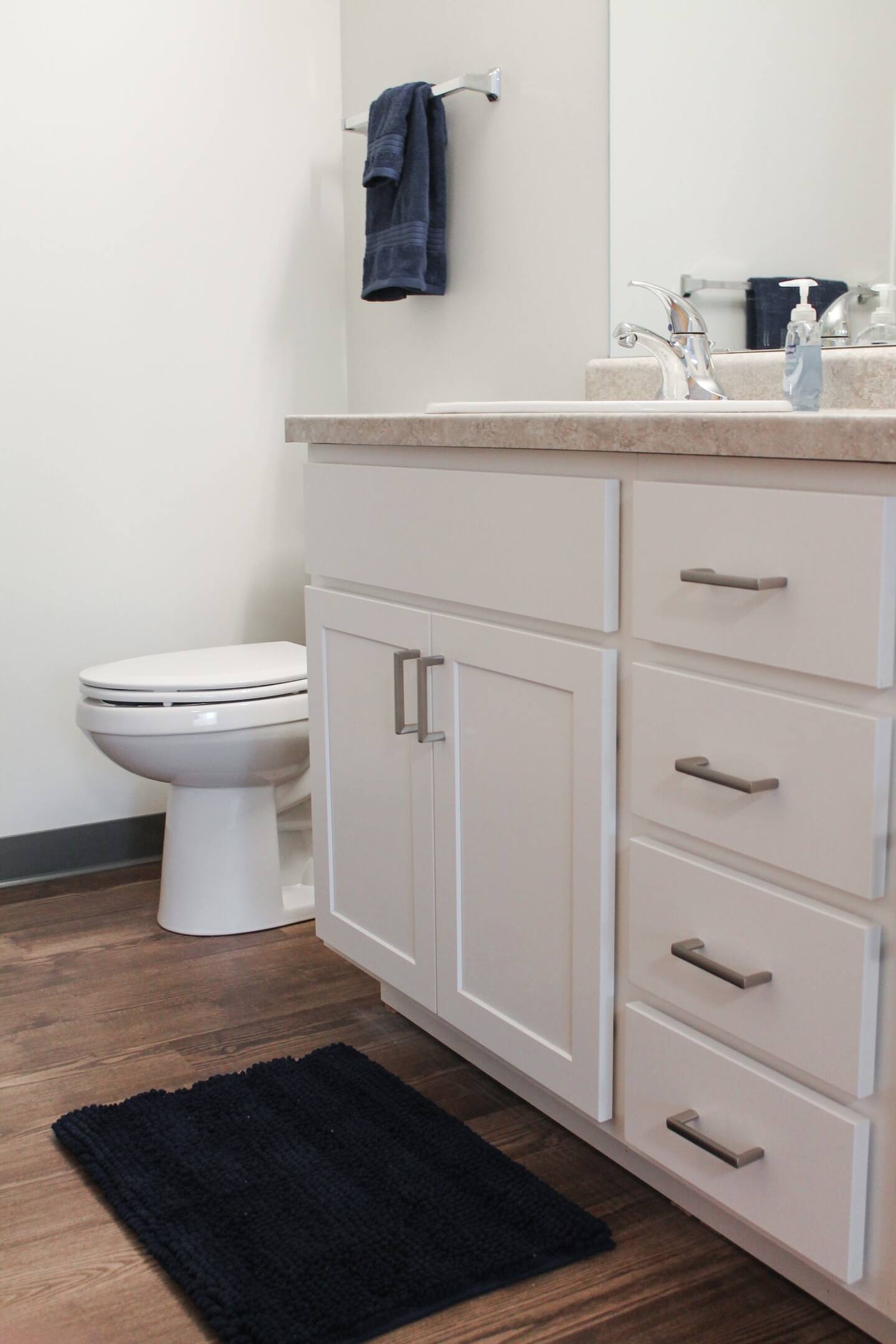 A bathroom with white cabinetry, a countertop, a sink, a toilet, a wall-mounted towel rack with a blue towel, a blue bath mat, and a hand soap dispenser on the counter.