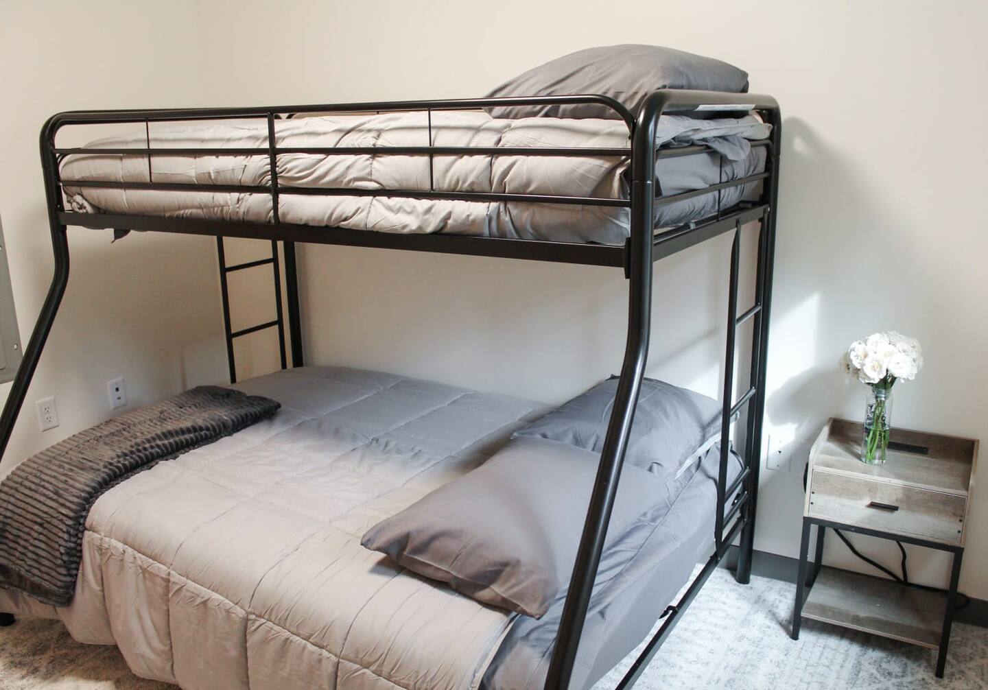 A neatly made bunk bed with black metal frame features a twin upper bed and a full lower bed with grey bedding. Next to the bed is a wooden nightstand holding a vase of white flowers.