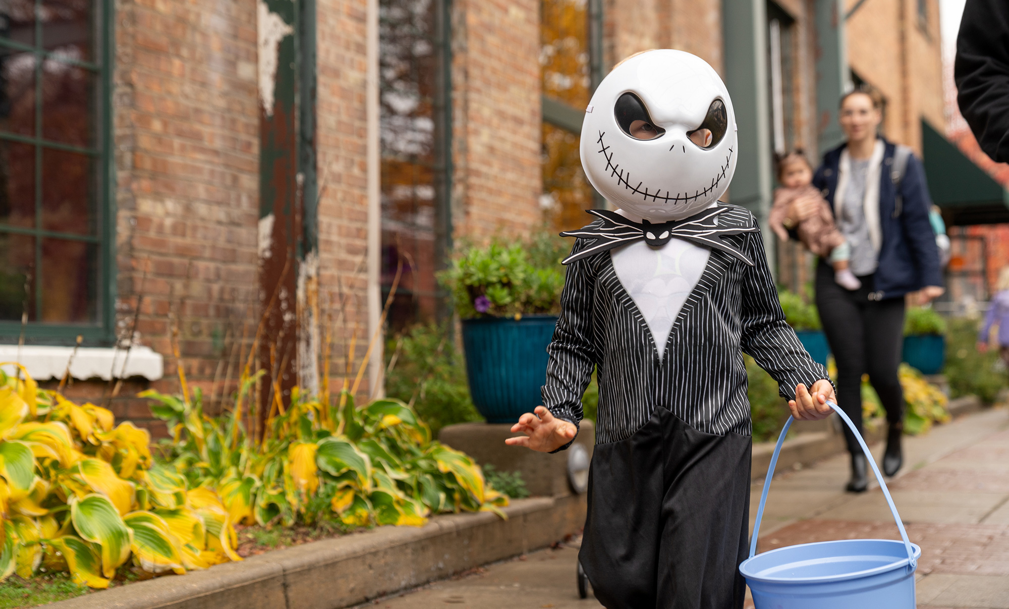 A child in a Jack Skellington costume carries a blue bucket while walking down a sidewalk. Plants and a brick building are in the background, with a person carrying a child following behind.