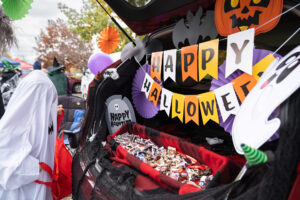 A trunk decorated with "Happy Halloween" banners, Halloween-themed decorations, and filled with candy. A person in a costume is visible on the left side of the image.