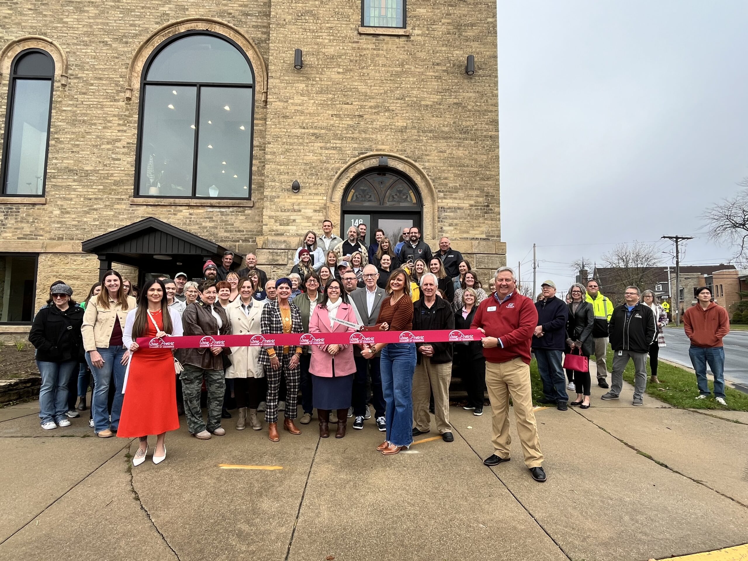 A group of people pose outside a large brick building for a ribbon-cutting ceremony. A red ribbon is held at the front, with several individuals standing closely behind it.