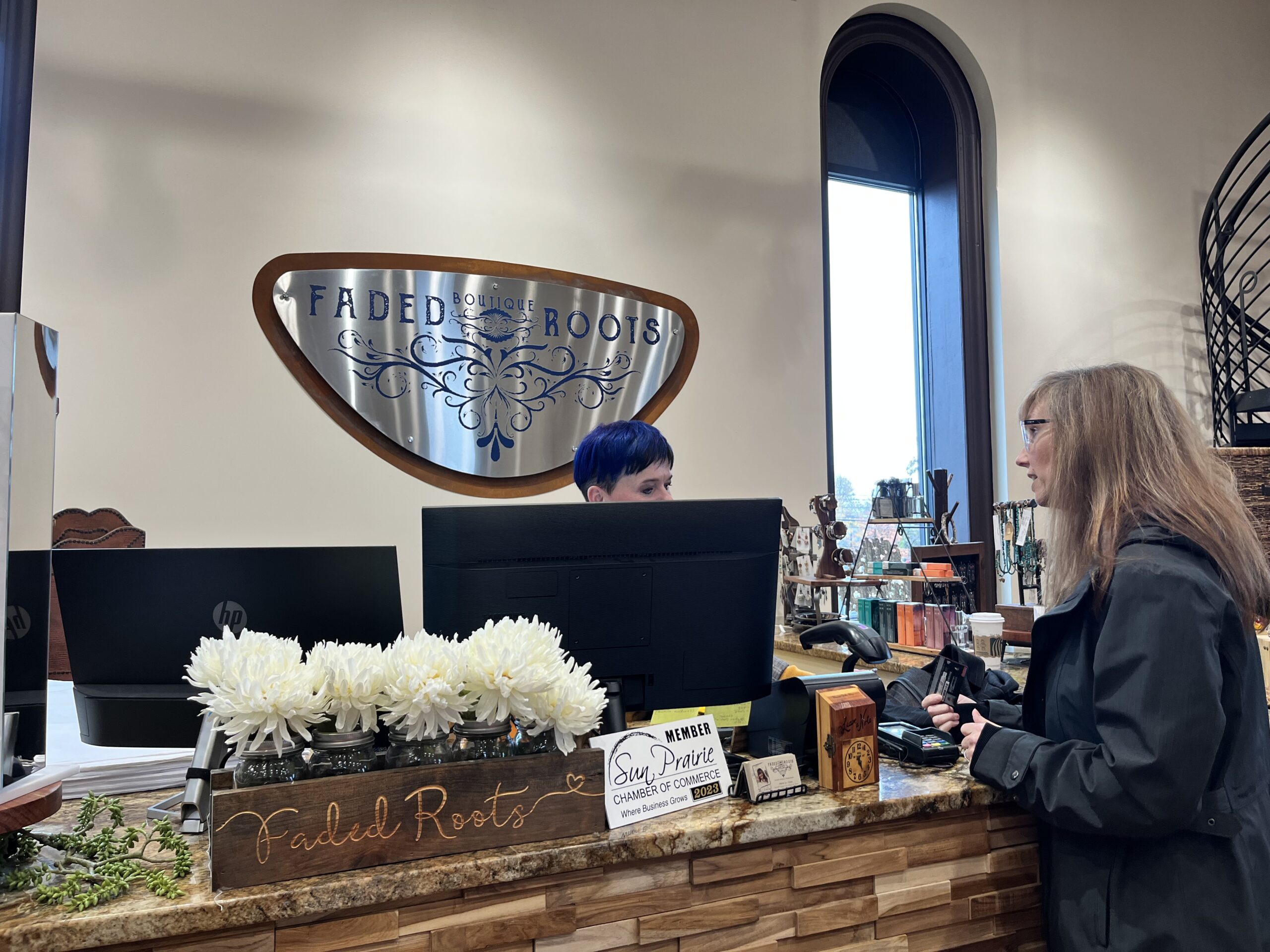 A woman with long hair speaks to a blue-haired store employee at the Faded Roots boutique counter, decorated with white flowers and a sign.