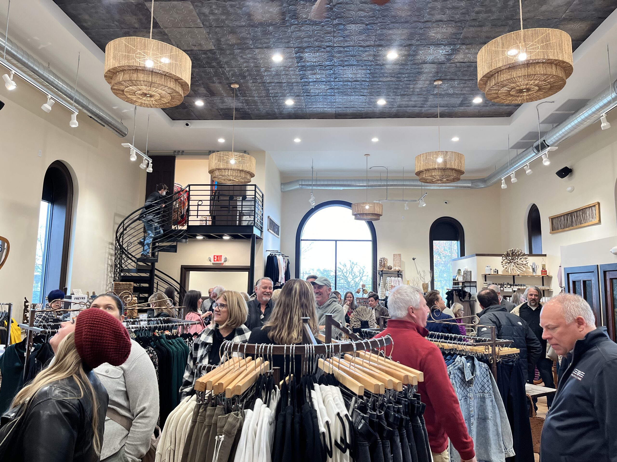 A busy clothing store interior with shoppers browsing racks of clothes, featuring a spiral staircase and large arched windows.