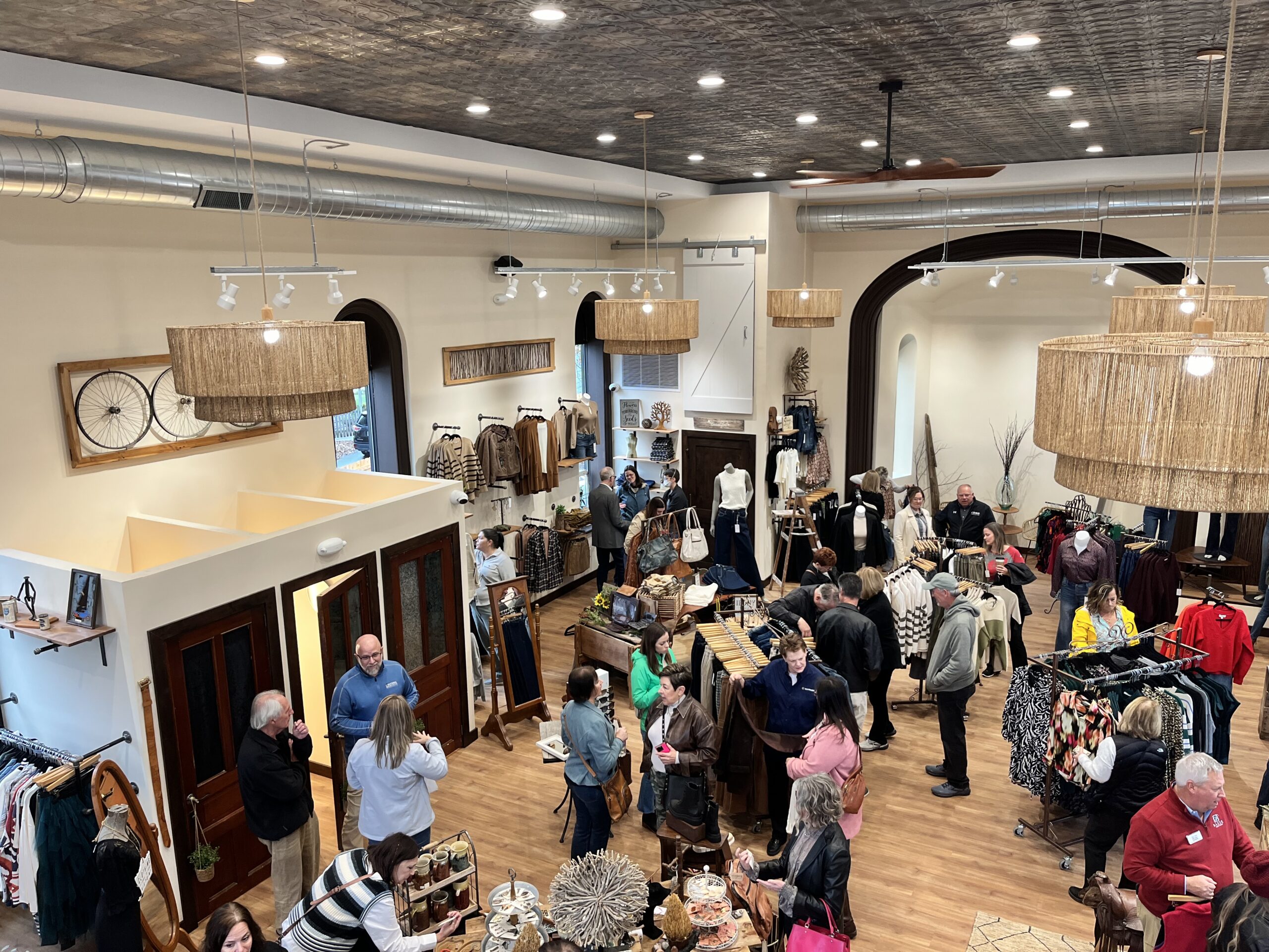 People shopping and interacting in a clothing store with racks of clothes, decorative lighting, and a bicycle on the wall.