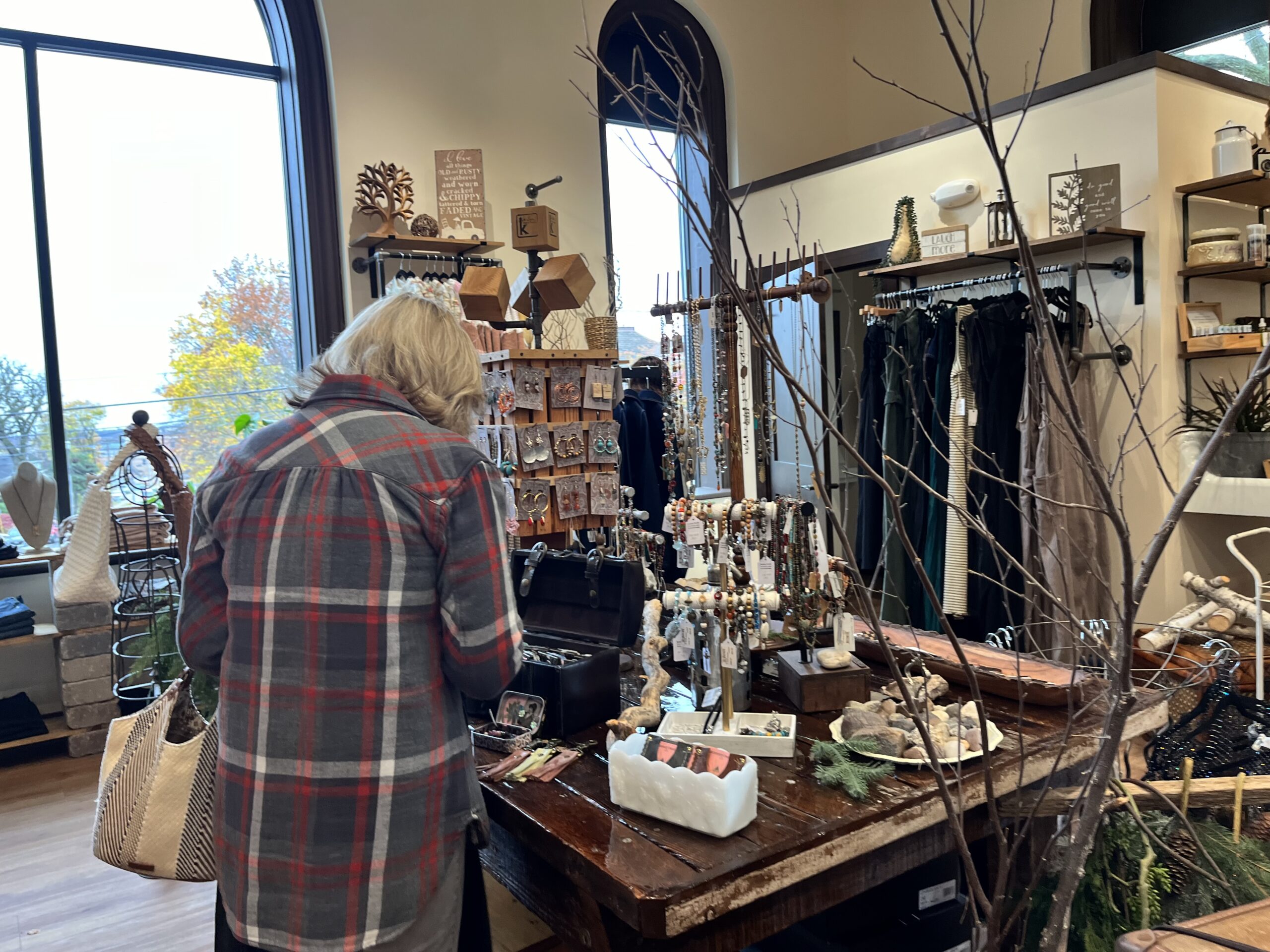 Person browsing goods in a rustic shop with various handmade items, jewelry, and decorations displayed on wooden tables. Large window in the background.