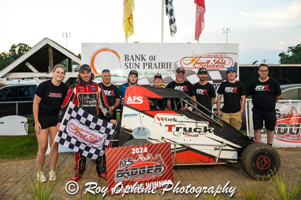 A race car driver in racing gear holds a checkered flag and stands with a team beside a small race car at Angell Park Speedway.