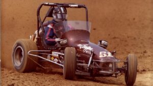 A person wearing a helmet drives a black sprint car with the number 51 on a dirt racetrack, with dust being kicked up by the tires.