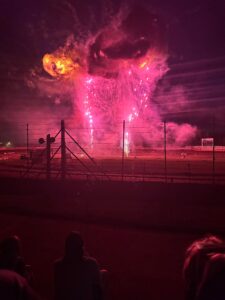 Bright pink fireworks and smoke illuminate the night sky over a fenced outdoor venue, with silhouetted spectators in the foreground.