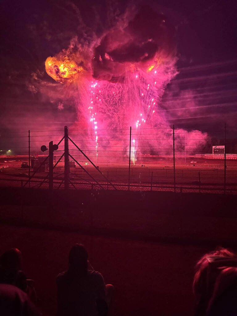 Bright pink fireworks and smoke illuminate the night sky over a fenced outdoor venue, with silhouetted spectators in the foreground.