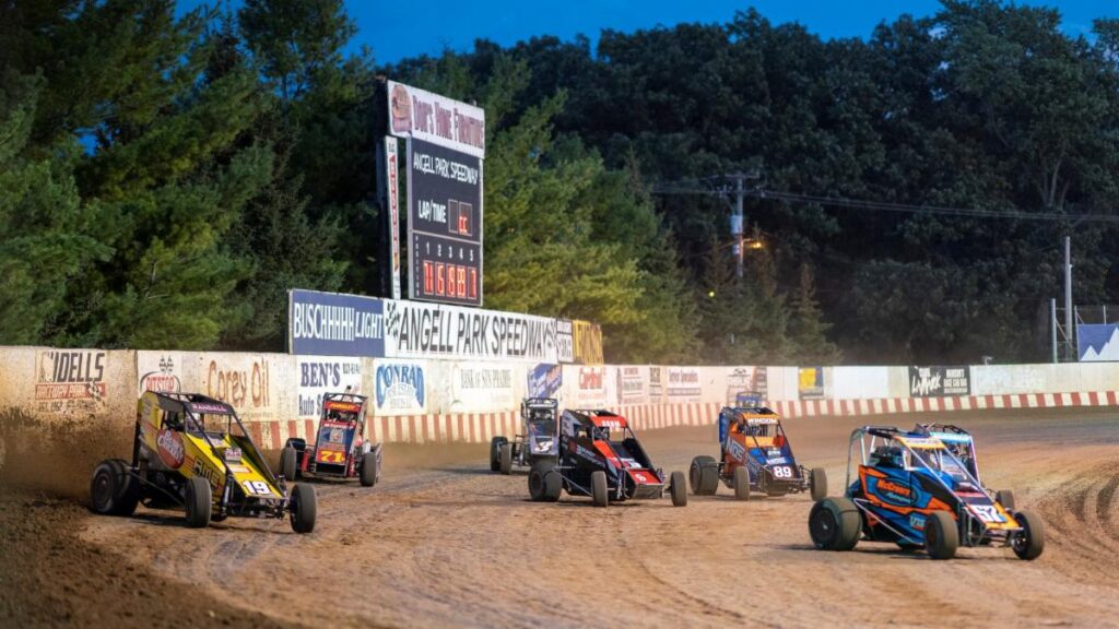 Race cars navigate a dirt track at dusk, with trees and a scoreboard in the background.
