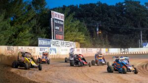 Race cars navigate a dirt track at dusk, with trees and a scoreboard in the background.