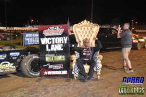 A race car driver sits on a gold throne-style chair, holding checkered flags, next to a "Victory Lane" sign at a dirt track with cars and people in the background.