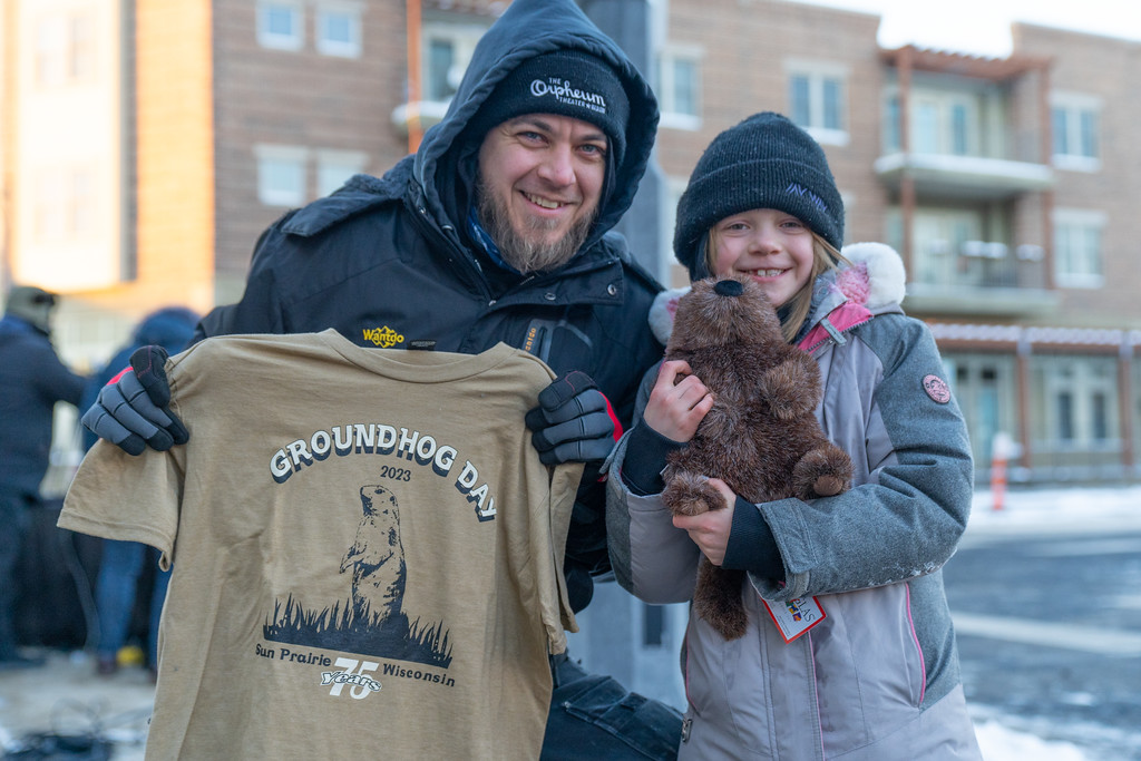 A man and a girl in winter clothing hold a Groundhog Day shirt and a plush toy outdoors.