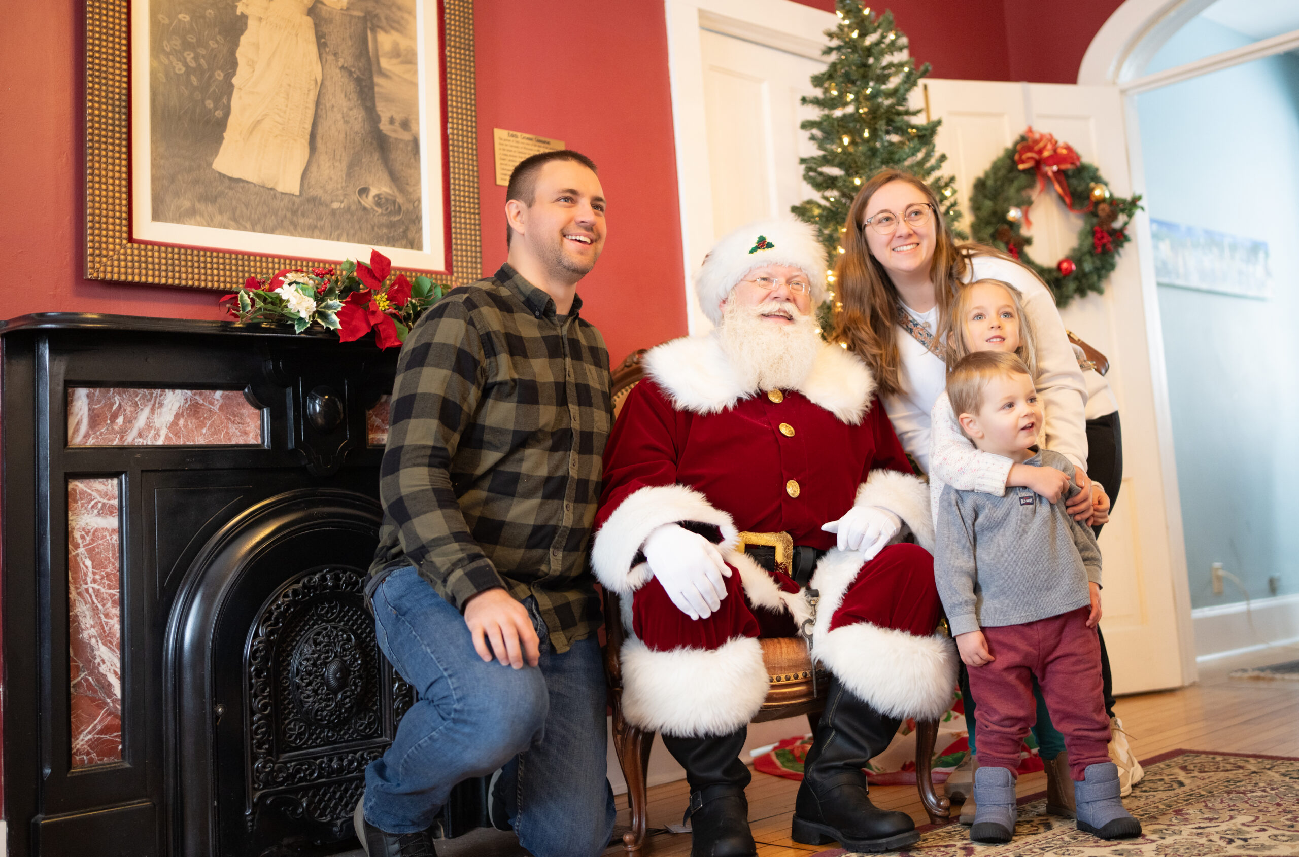 A family poses next to Santa Claus in a festive room decorated with a Christmas tree, wreath, and poinsettias.