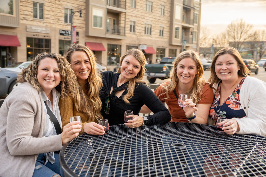 Five women sitting at an outdoor table, smiling and holding small glasses, with buildings in the background.
