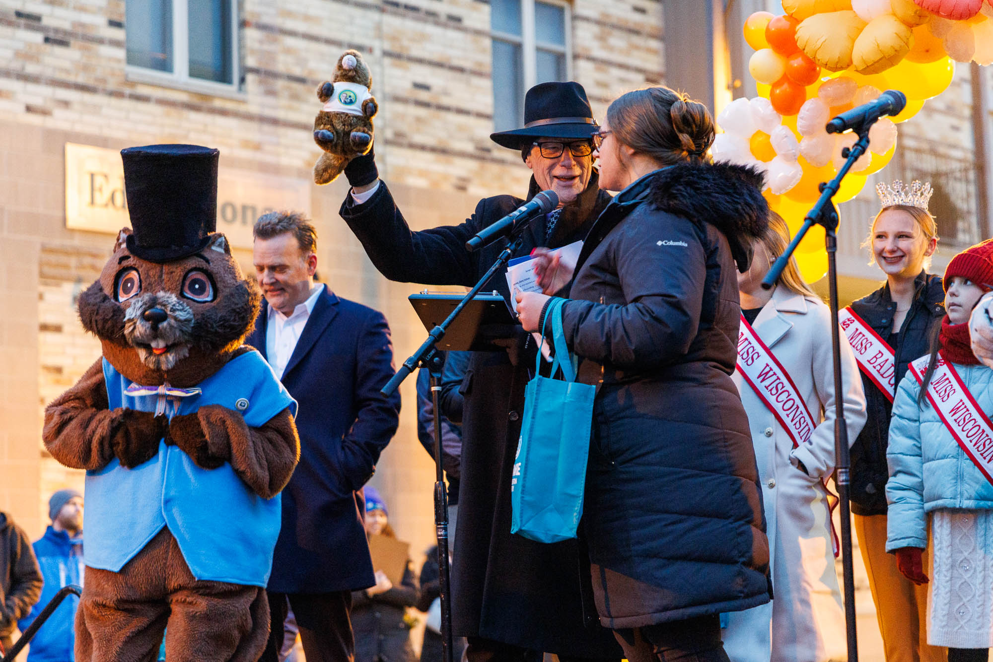 A man in a suit holds a stuffed animal next to a person in a groundhog costume. A woman with a microphone stands nearby. Balloon decorations and people in pageant sashes are in the background.
