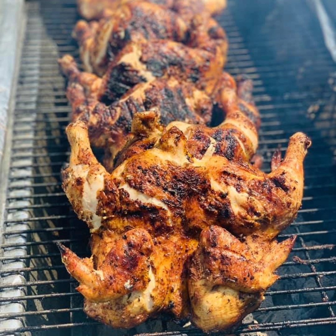 Several whole chickens are being grilled on a barbecue, seasoned and cooked over open flames with visible grill marks.