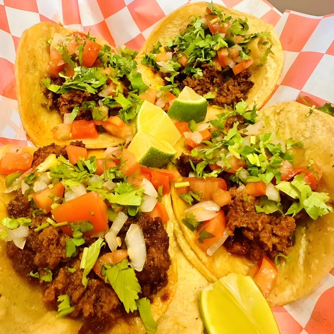 Four tacos topped with seasoned ground meat, diced tomatoes, onions, and cilantro, served on a red and white checkered paper with lime wedges.