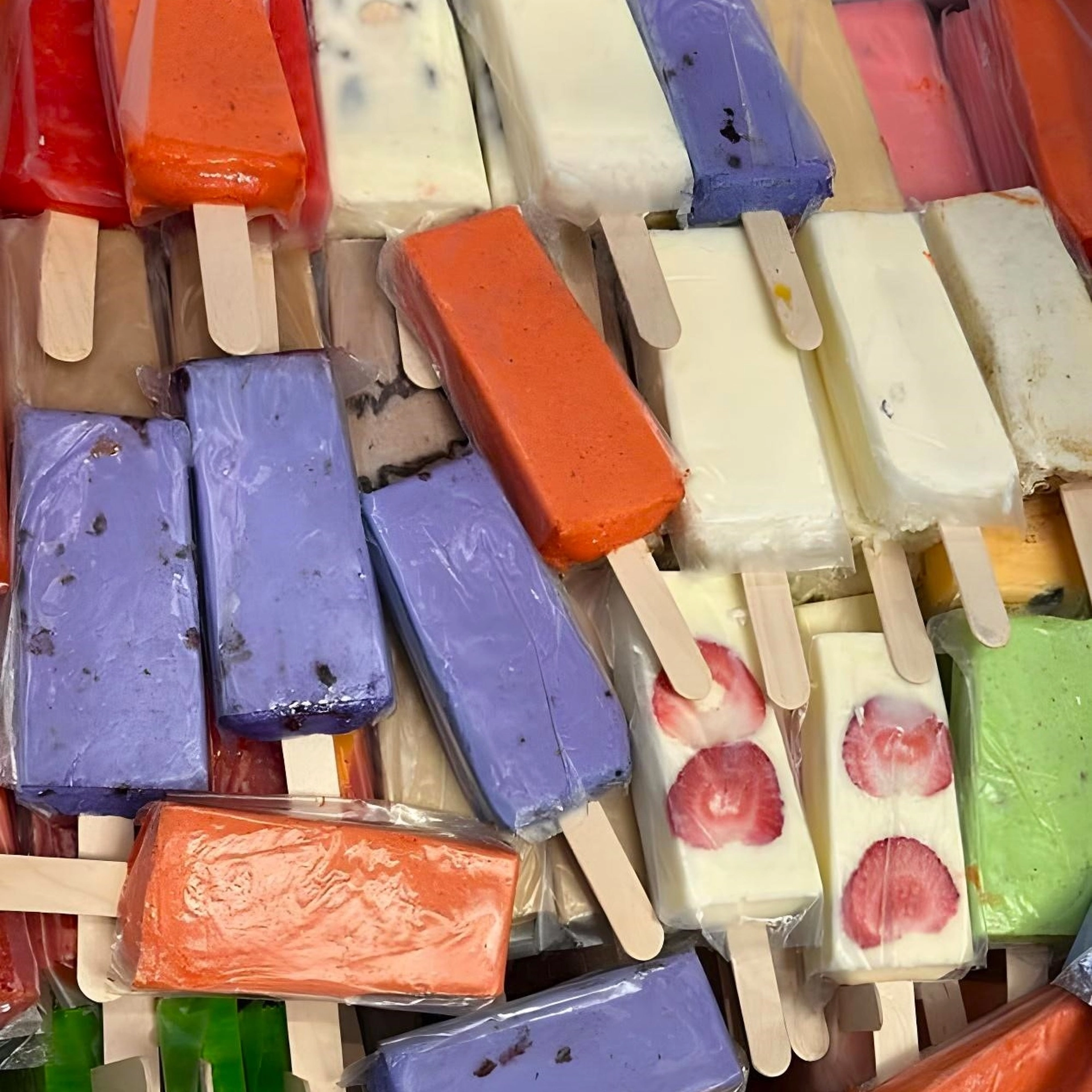 A variety of colorful, rectangular popsicles with different flavors, some containing visible fruit pieces, displayed closely together on wooden sticks.