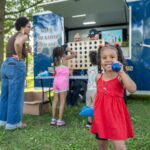 A young girl in a red dress smiles while holding a blue ball at an outdoor event near a police truck, with children and adults interacting in the background.