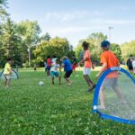 Children play soccer on a grassy field with small portable goals, surrounded by trees and people on a sunny day.