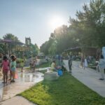 People gather at a park near a playground and splash pad on a sunny day; a trailer labeled "Go-Cart" is parked nearby and children play in the water.
