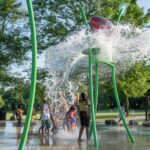 Children play under a large splash of water from a green structure at a splash pad in a sunny park, surrounded by trees and grass.