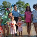 A group of six people, including four children and two adults, stand and pose together in a splash pad park on a sunny day.