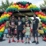 Five people, including a child holding a basketball, stand under a red, yellow, green, and black balloon arch at an outdoor event in a park.