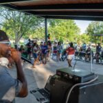 A DJ performs with a microphone while a group of people, including children, gather and socialize under a park pavilion during a daytime outdoor event.