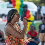 A woman with braided hair sings into a microphone at an outdoor event, with people and colorful red, yellow, green, and black balloons in the background.