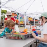 Children and adults sit at a table under white canopy tents doing arts and crafts with colorful materials at an outdoor event.