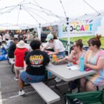 People sit at picnic tables under white tents, participating in arts and crafts activities at an outdoor event next to a colorful trailer.