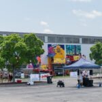 Artists work on colorful murals on the side of a large building, with lifts, tents, and a painted van visible in a parking lot.