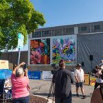 People gather outdoors near a building with two large colorful murals depicting flowers and abstract designs; some are photographing or filming the scene.
