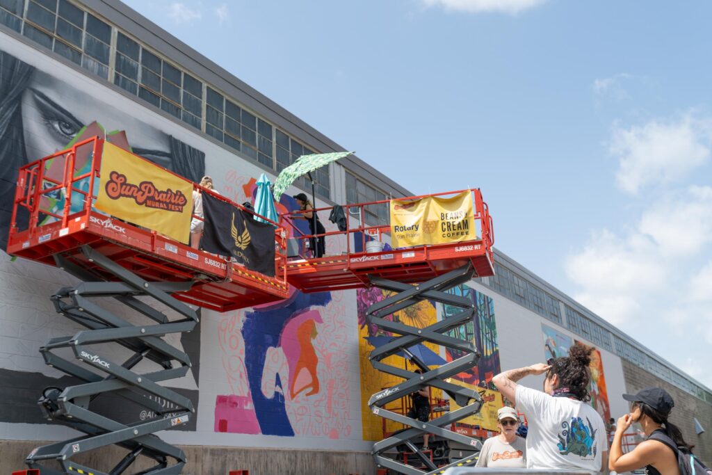 Artists on scissor lifts paint large murals on a building, while several people watch and take photos from the ground under a sunny sky.