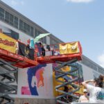 Artists on scissor lifts paint large murals on a building, while several people watch and take photos from the ground under a sunny sky.