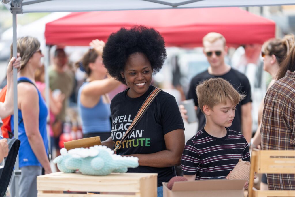 A woman and a boy stand at a market stall with handmade items, surrounded by other people under a canopy. The woman is smiling and holding a clipboard.